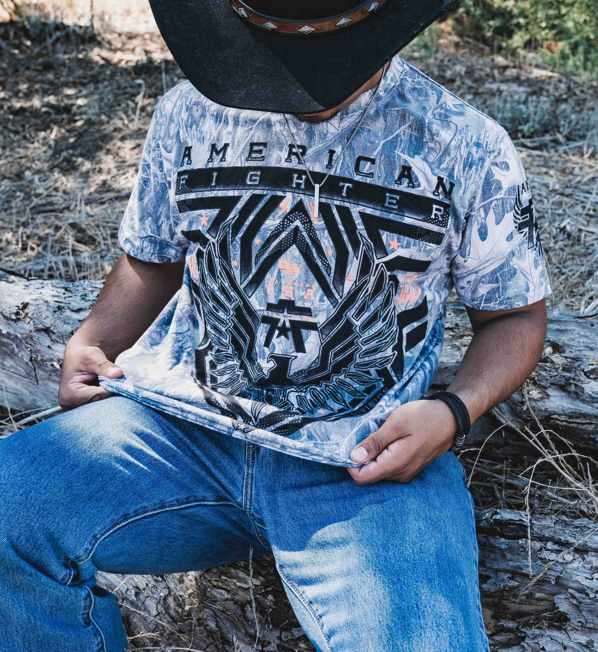 Person wearing an 'American Fighter' t-shirt sitting outdoors on a log.