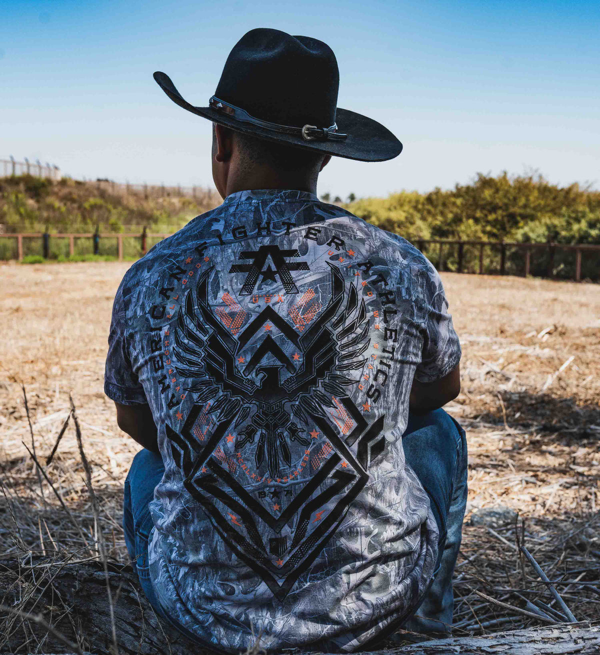 Person wearing a patterned shirt with visible branding, sitting in an open field.