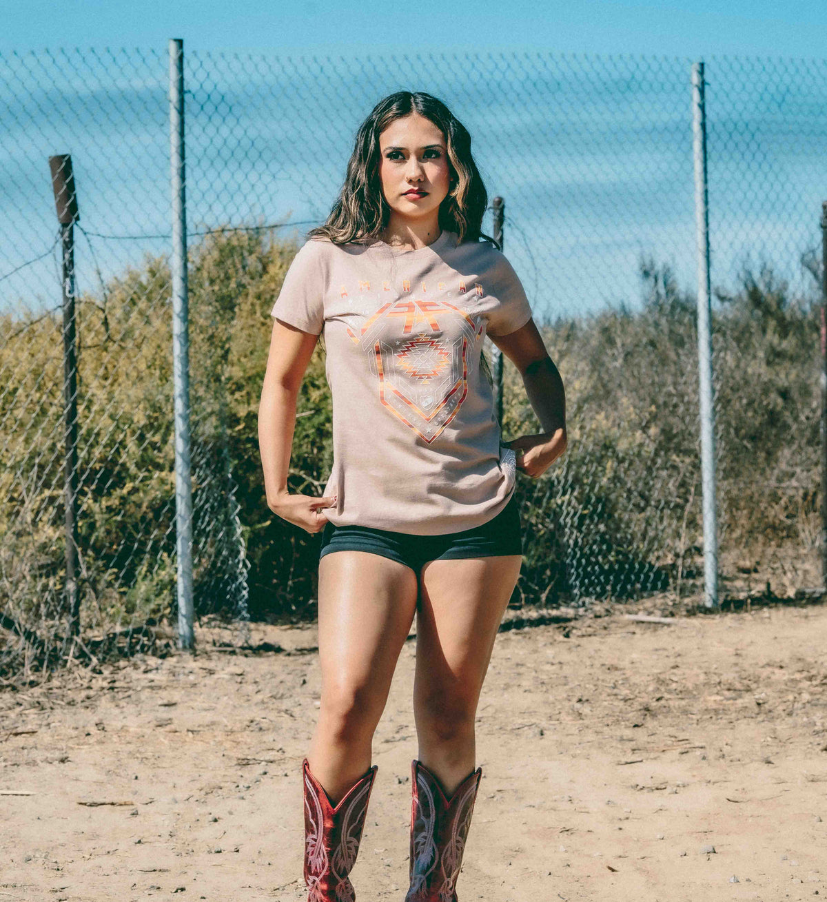 Woman wearing a graphic t-shirt and cowboy boots standing in an outdoor setting with a fence and trees in the background.