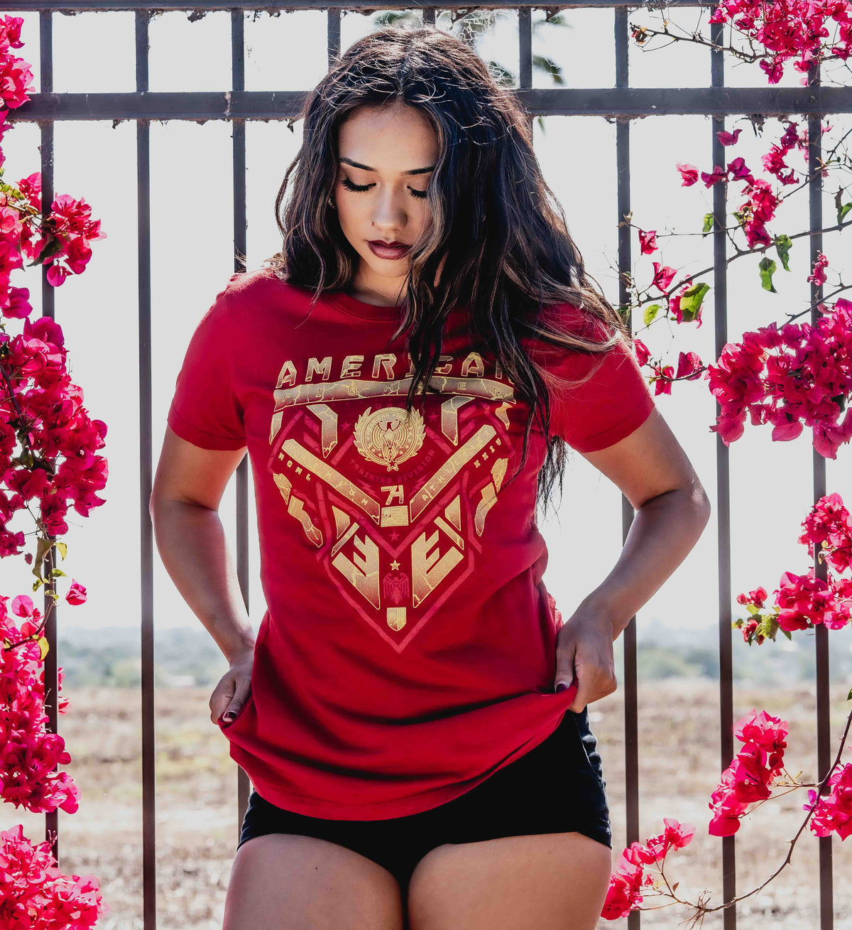 Woman wearing a red t-shirt with a gold graphic design, standing in front of a floral background.