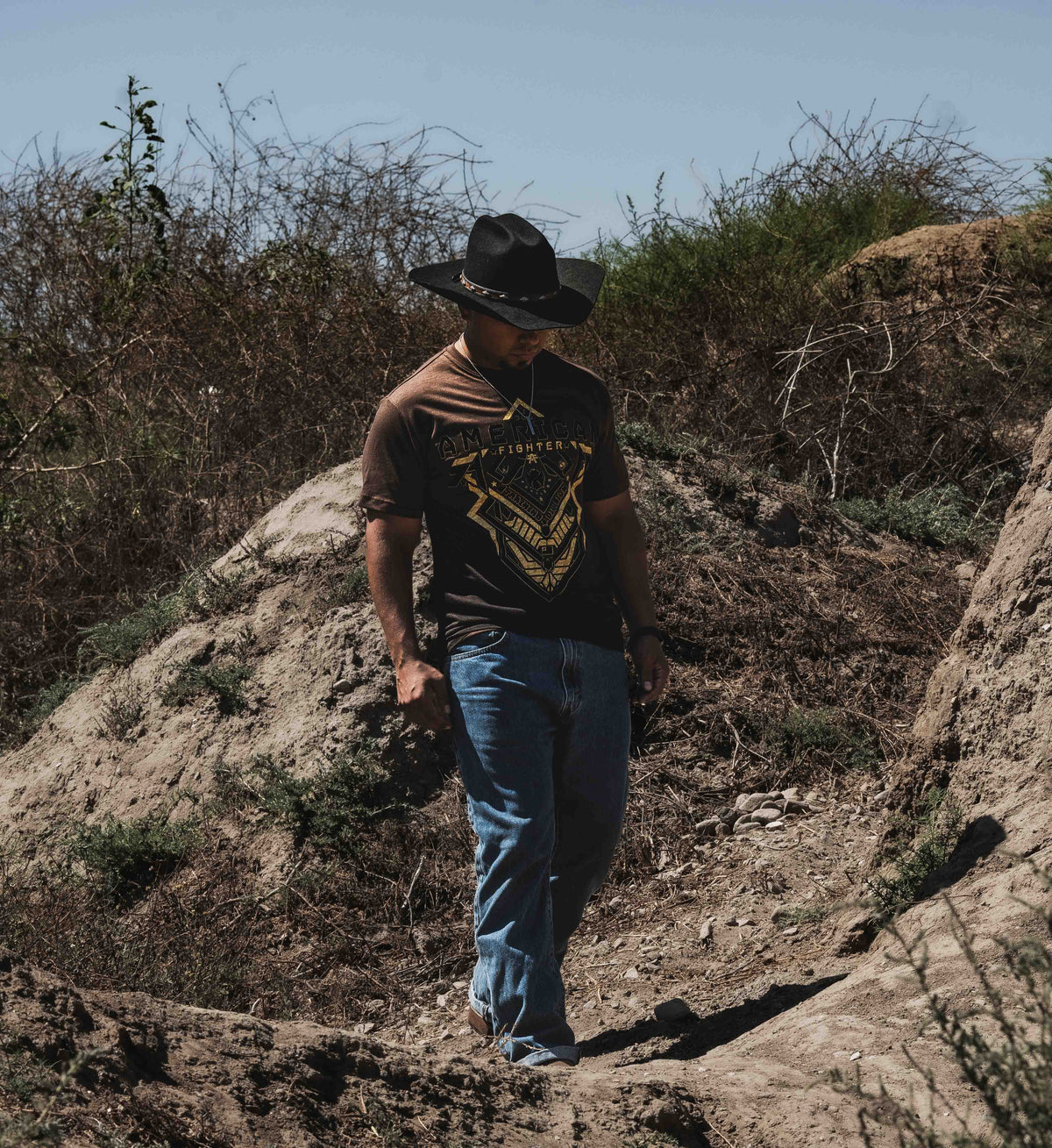 Man wearing a cowboy hat and graphic t-shirt walking on a rocky trail with dry vegetation.
