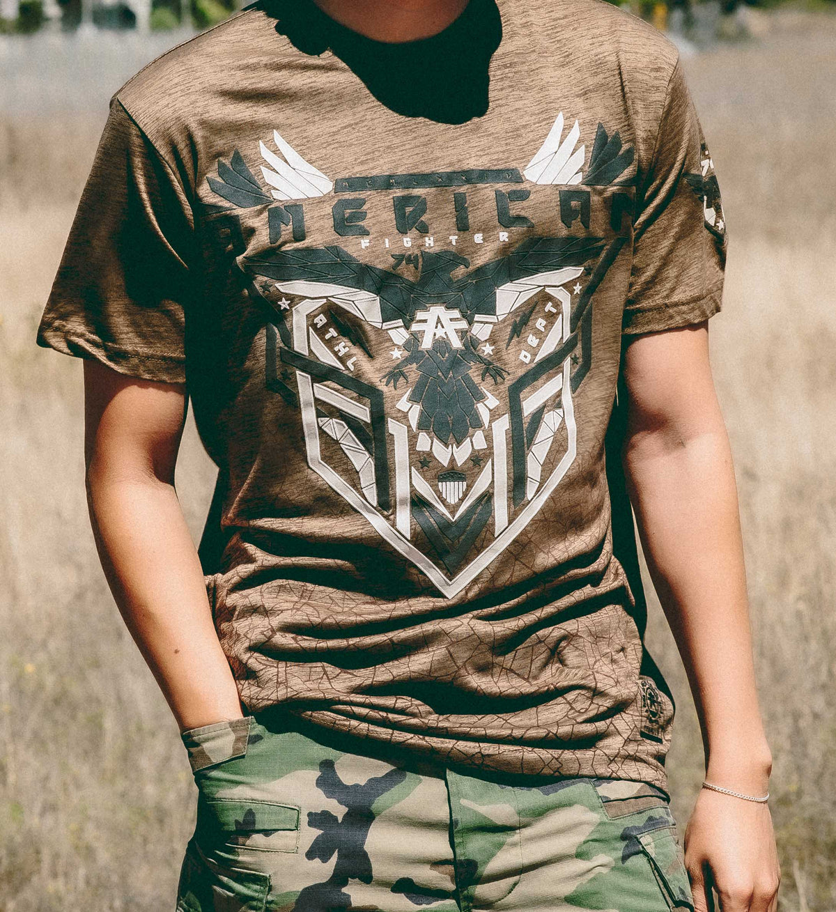 Person wearing a brown t-shirt with a graphic design and camouflage pants outdoors.