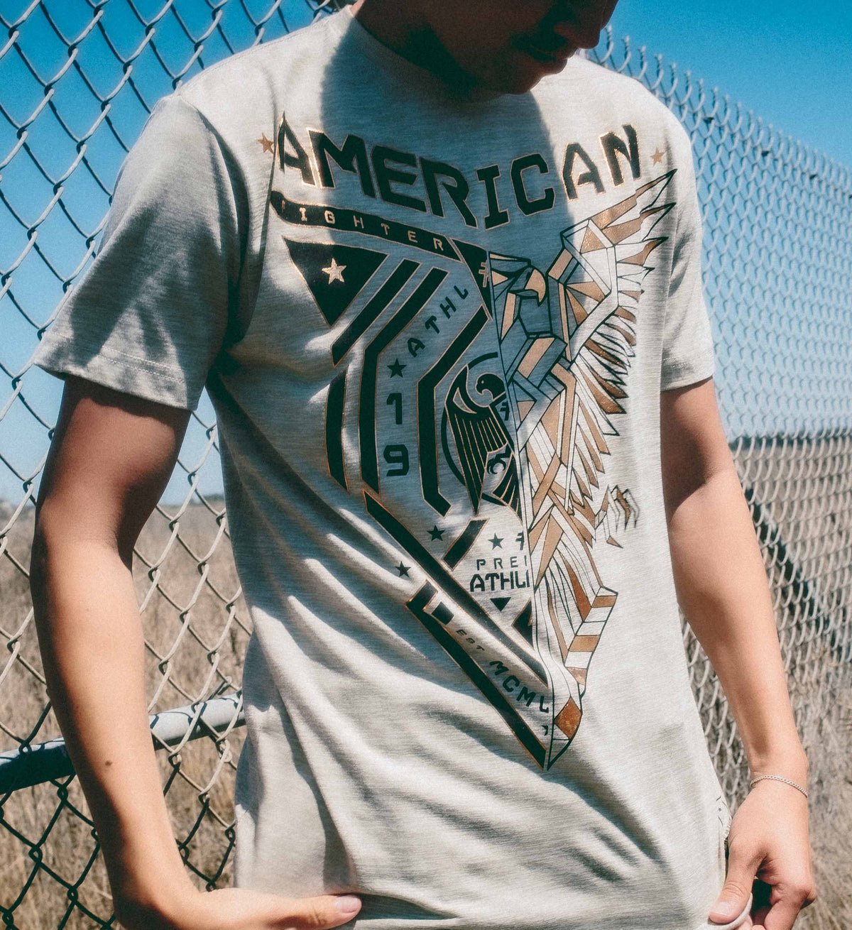 Person wearing a gray t-shirt with a graphic design in front of a chain-link fence.
