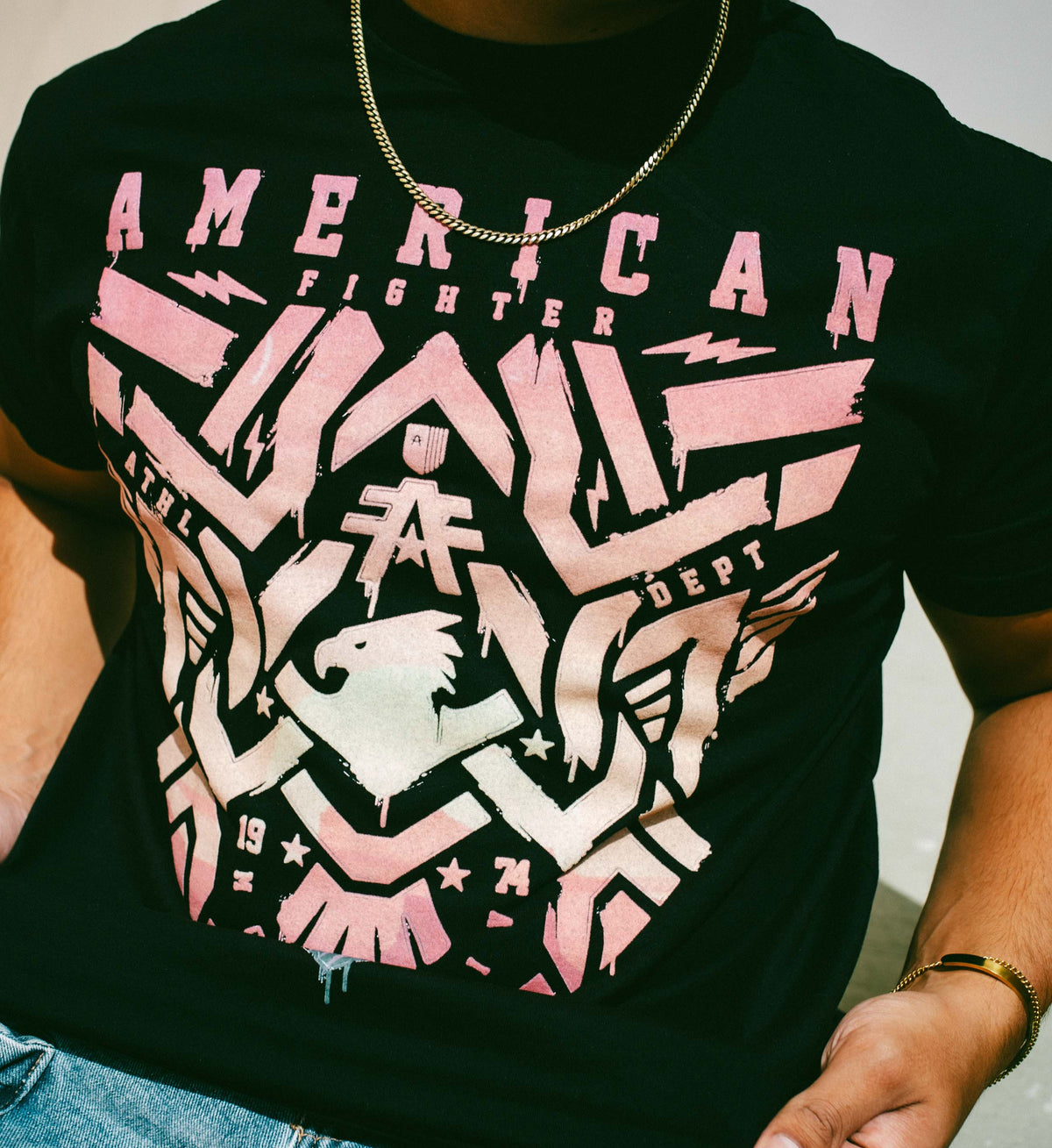 Person wearing a black t-shirt with pink and white graphic design, featuring the brand 'American Fighter'.
