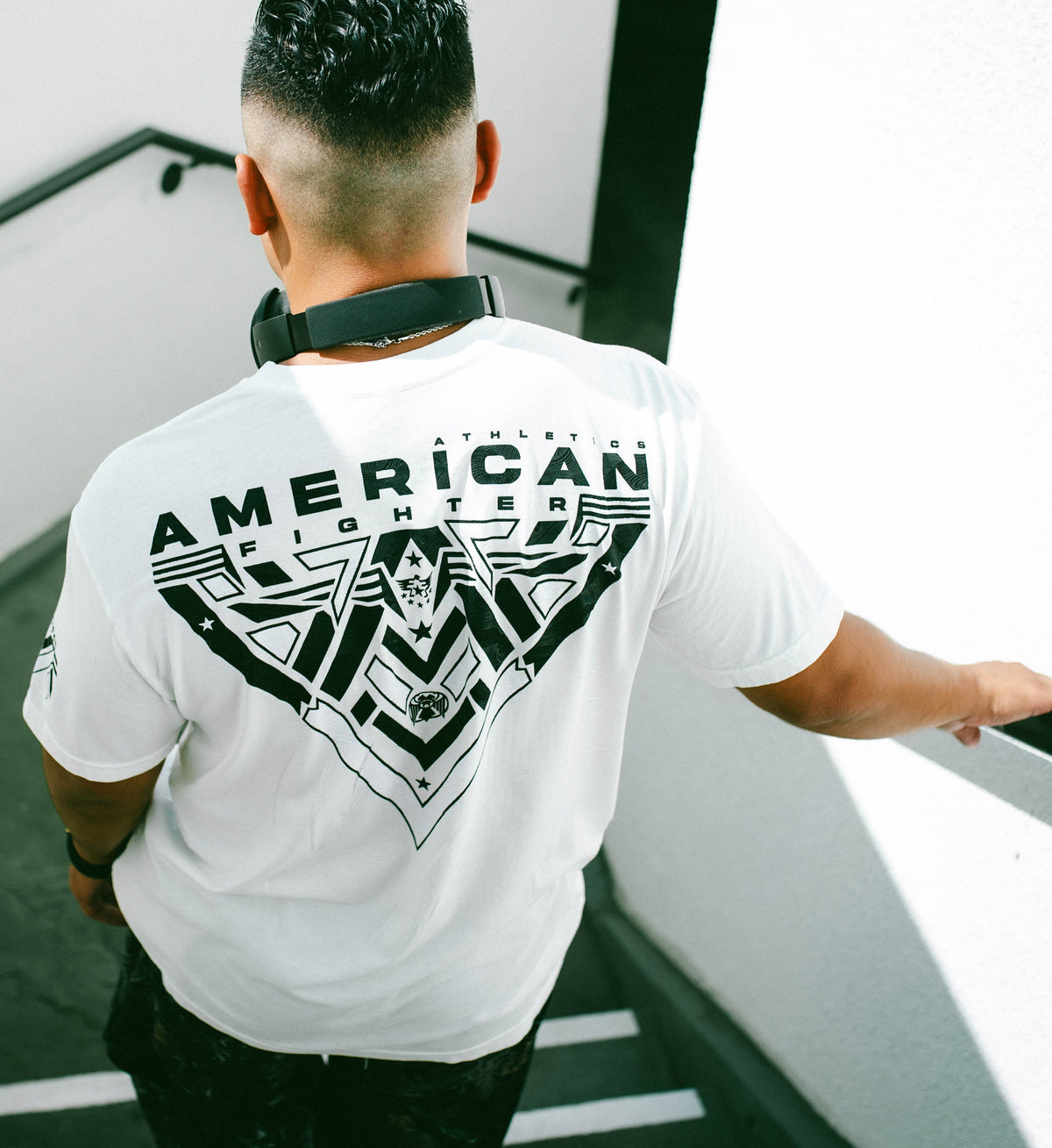 Person wearing a white 'American Fighter' t-shirt with a black graphic design on the back, standing in an indoor setting.